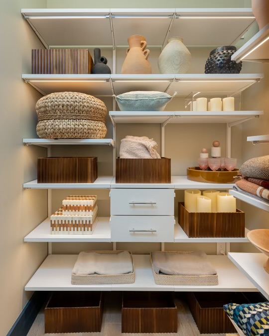 A storage closet after organizing: white shelves with soft under-shelf lighting, woven baskets, wooden bins, folded throws, candles and ceramic vases, with two white drawers in the center