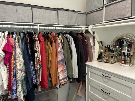 A walk-in closet with clothes hung by type, labelled storage boxes on the top shelf and a white dresser